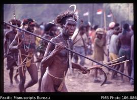 [Man holds long fighting spear; bib, feather headdress; in left hand he holds arrow shafts (?) an...
