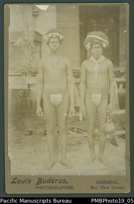 Mounted photograph of two Papua New Guinean men standing in front of a dwelling.
