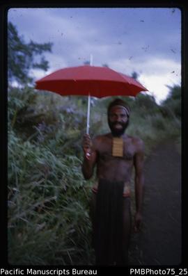 NG [New Guinea] Mt Hagen [man posing with a smile, in traditional clothing holding an open red Eu...