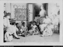 Group of women, men, and children sitting with Conrad and Christina Stallan inside mission house,...