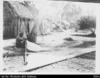 Man with hat sitting on ground in front of thatch buildings, working on a canoe sail (Duplicate o...