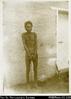 Solomon Islands man, with legs and hands bound together with chains, standing in front of wall. I...