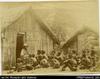 Solomon Islands men and boys sitting on ground in front of dwelling with Woodford seated on chair...