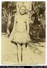 Portrait of girl in grass skirt standing against tree-trunk. Written on back in pencil: 'Woman of...