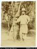 Portrait of Woodford holding a rifle, standing by a palm tree.