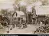 View of village with women seated on ground in clearing in front of dwellings built on stilts. Mo...
