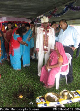 [Suva Wedding  Mahen the groom standing near his seated mother.  Candle in grass has been lit. ]