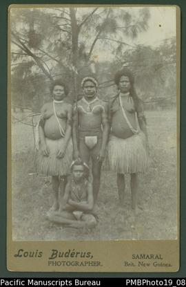Mounted photograph of Papua New Guinean group. Two women and one man standing, with a child seate...