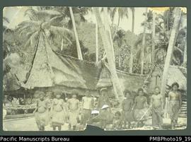 Ten people standing in foreground of Papua New Guinean village, canoe and coconut trees in backgr...