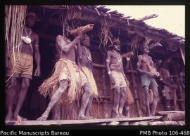 [Asmat men on front porch of longhouse; one blows in bamboo trumpet]