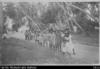 Village scene with line of Cook Islander women in dresses and hats walking down dirt road
