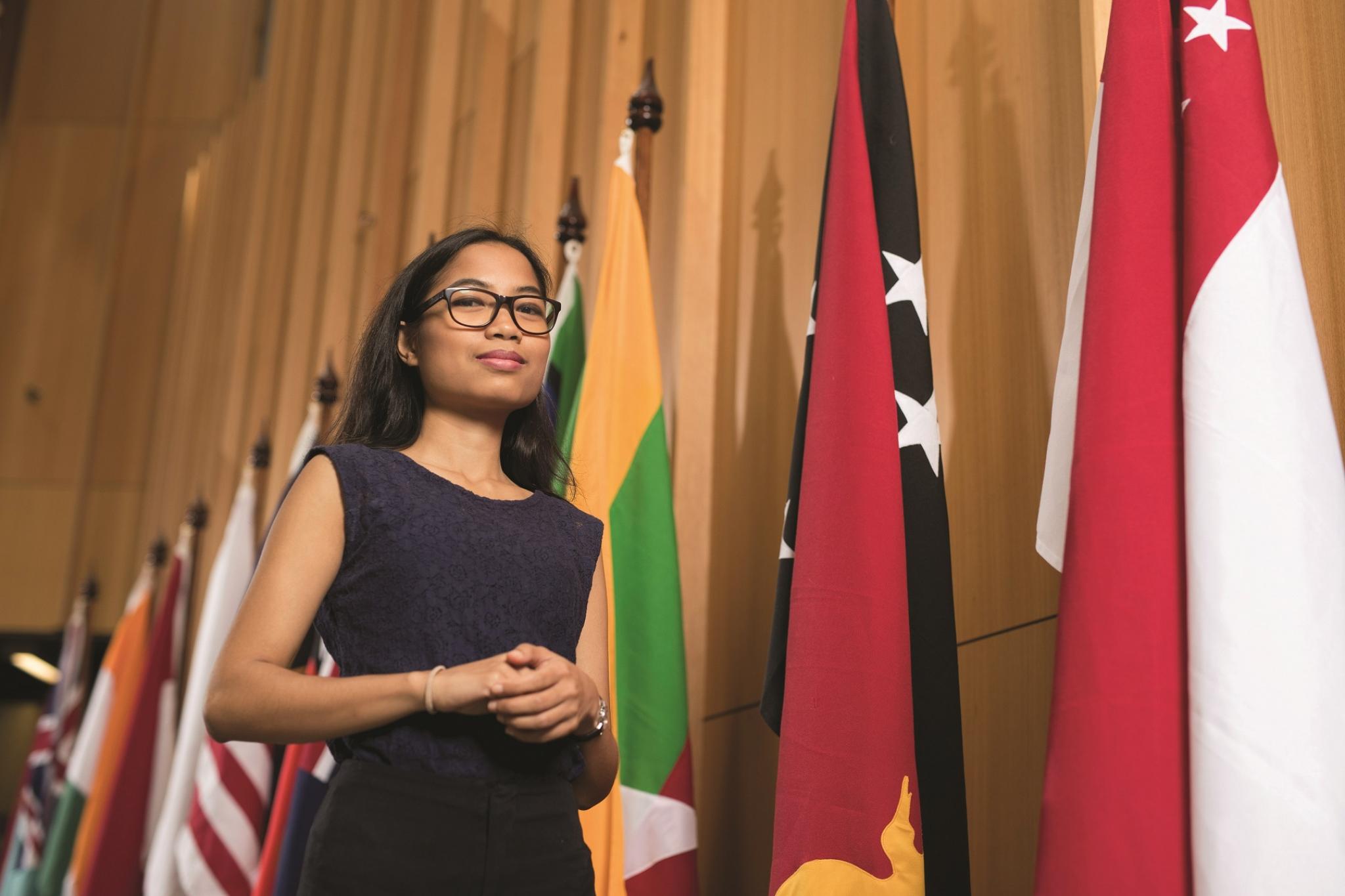 Woman in black dress with black glasses smiling at camera an standing in front of world flags
